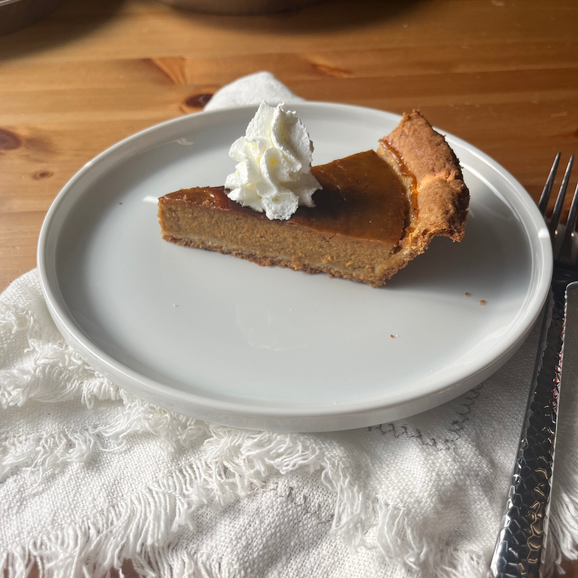 Slice of pumpkin pie with whipped cream on a white plate, placed on a wooden table.
