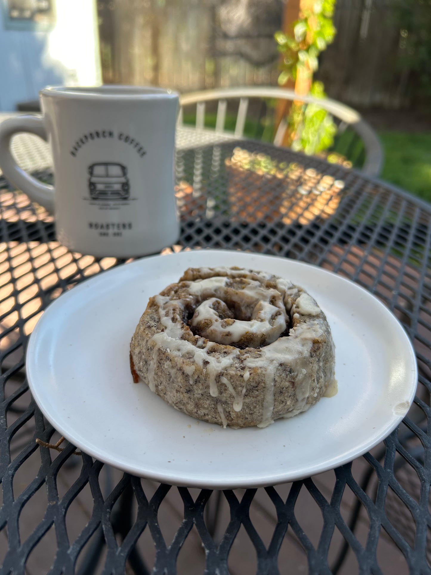 A cinnamon roll with white icing sits on a white plate on an outdoor metal patio table, with a white ceramic mug from Backporch Coffee Roasters in the background.