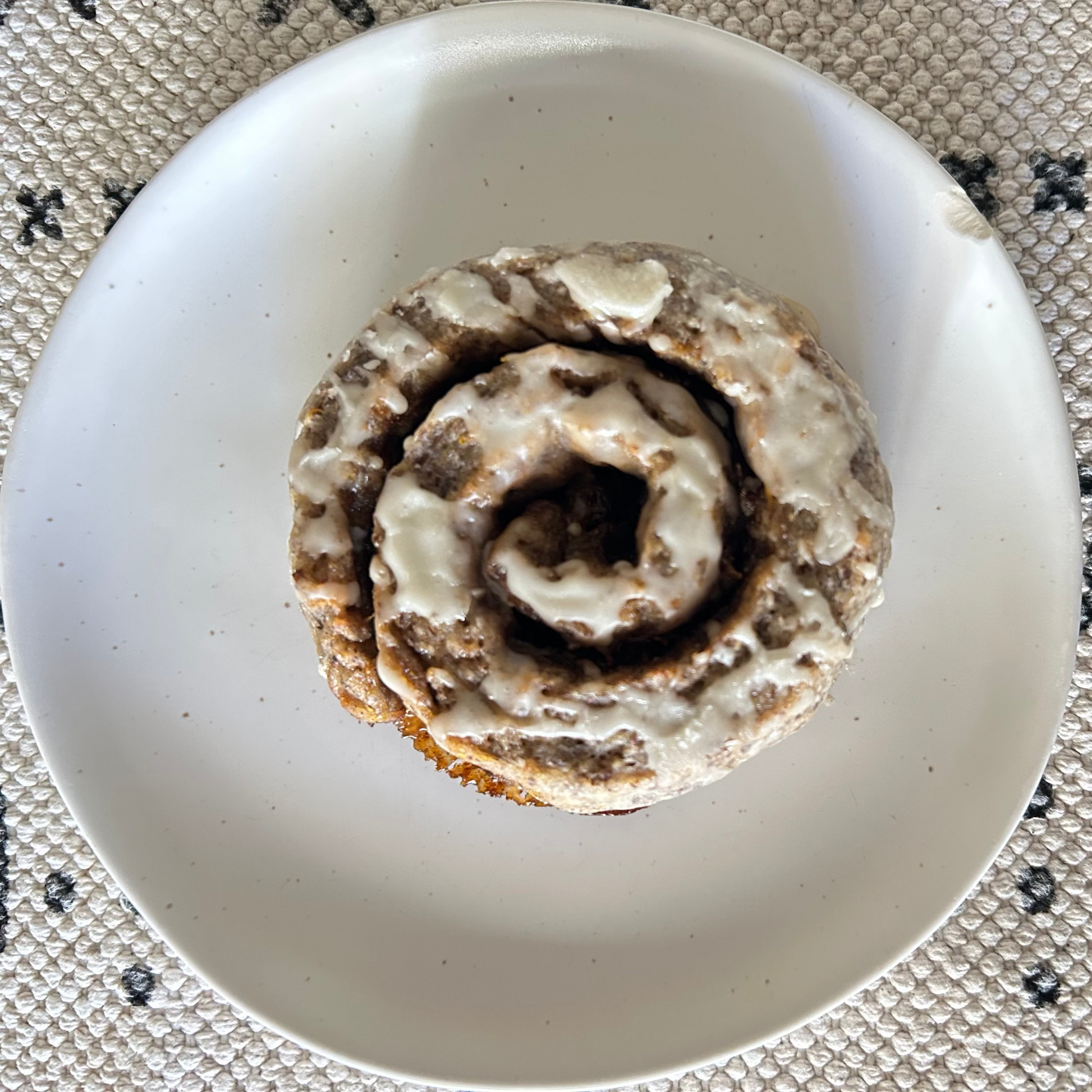 A single cardamom bun with white icing sits on a white plate, placed on a textured beige woven placemat with dark geometric patterns.