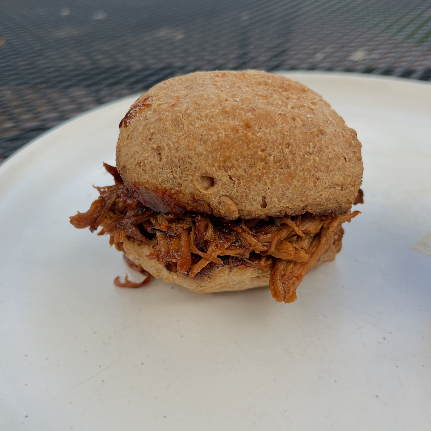 Pulled pork sandwich on a bun on a white plate with a blurred outdoor background