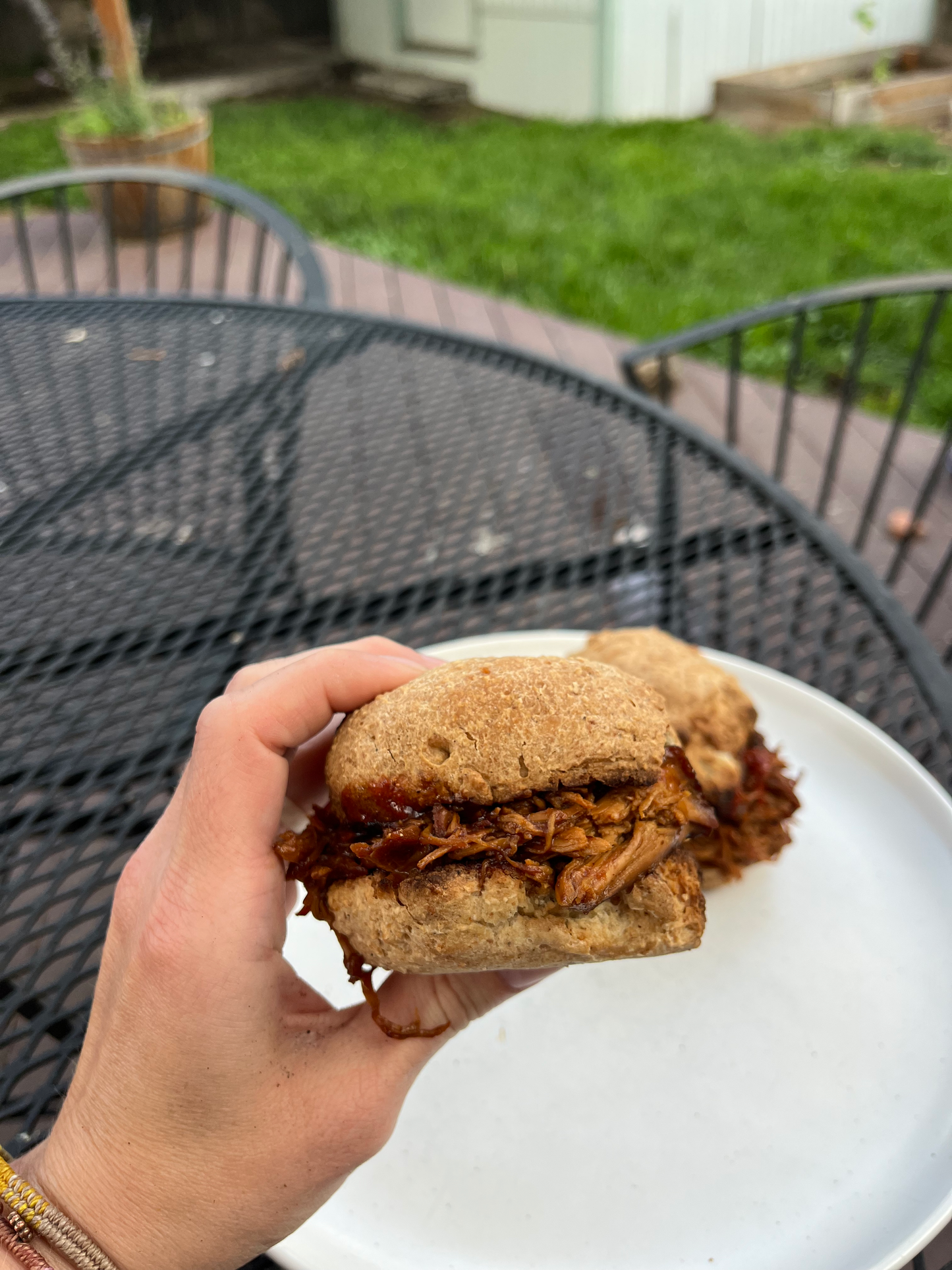 Hand holding a pulled pork sandwich on a plate with another sandwich in the background, outdoors.